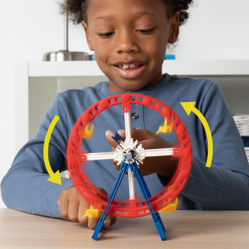 15266_Image3 A smiling child wearing a blue long-sleeve shirt is playing with a small model Ferris wheel. The Ferris wheel features red, blue, and white components and is positioned on a table. Yellow arrows are shown indicating the rotation direction of the Ferris wheel.